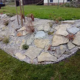 Rabata z kamieni i grysu w Lipnicy Wielkiej, z młodymi drzewkami i krzewami, obrzeże z trawnika.