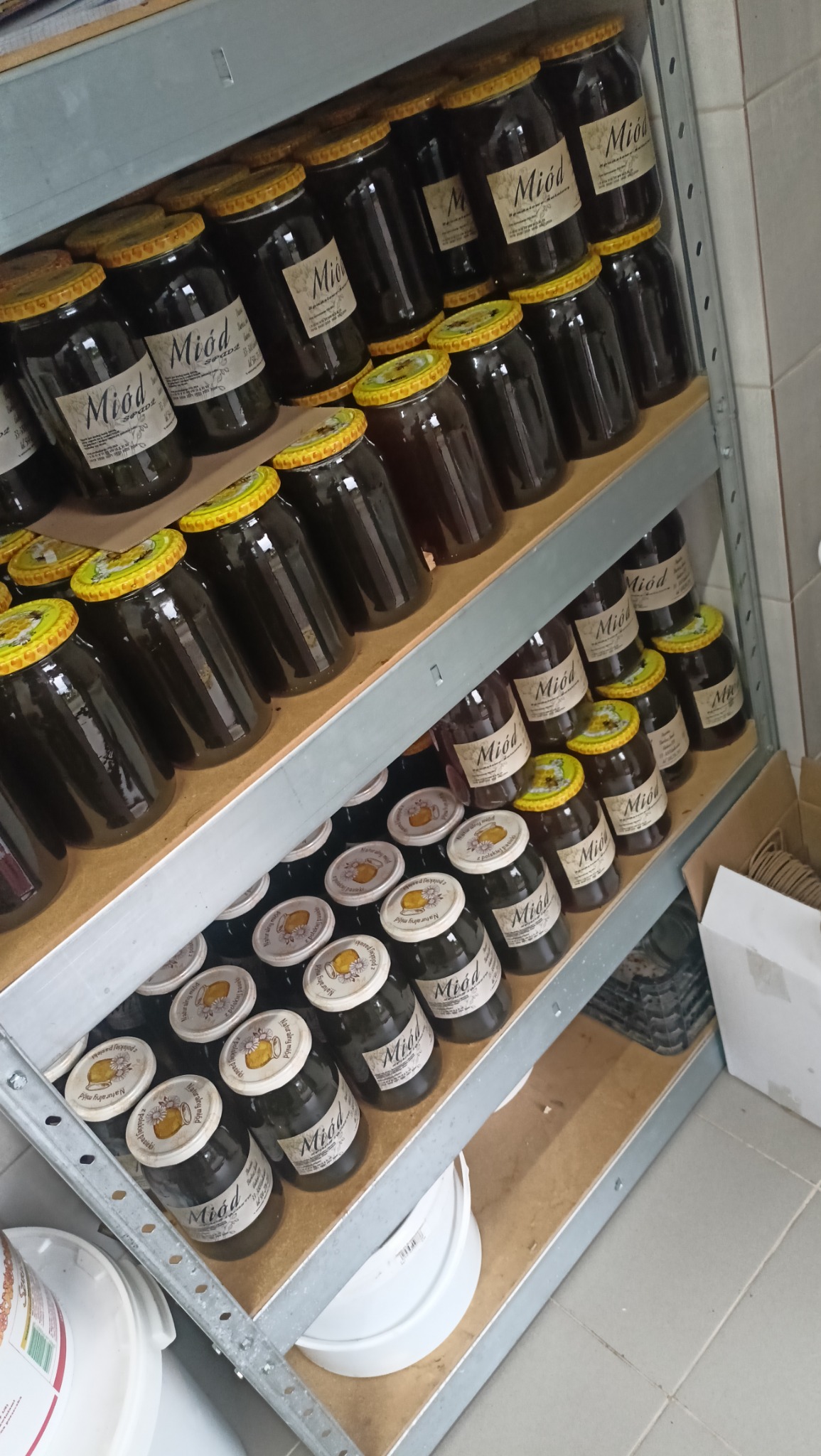 Półki regału wypełnione słoikami z ciemnym miodem, etykiety z napisem 'Miód' w języku polskim, różne wzory wieczek, widok z boku, oświetlenie sztuczne.