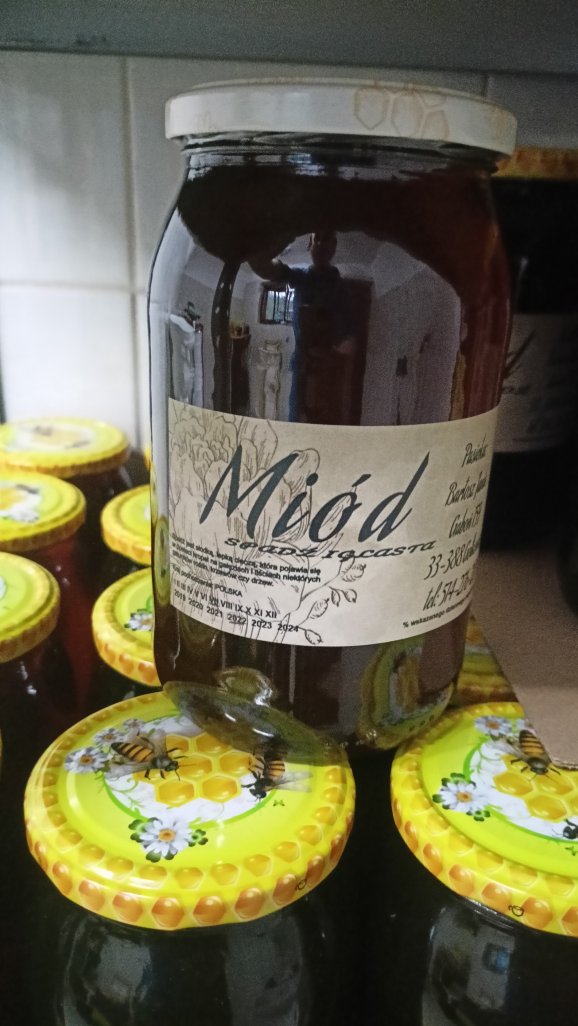 Słoik ciemnego nektaru spadziowego z etykietą i dekoracyjną nakrętką z motywem pszczół, otoczony innymi słoikami, widoczne odbicie osoby w szkle słoika.
