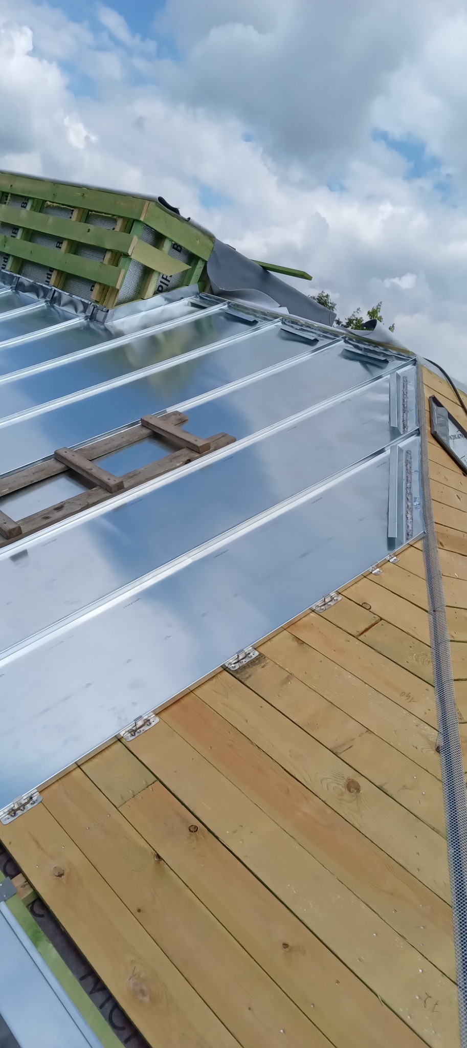 Montaż blachy na rąbek stojący na drewnianym poszyciu dachu, widoczne elementy mocujące i rusztowanie dachowe, pochmurne niebo w tle.