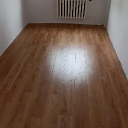 Nowo ułożone panele podłogowe o wzorze drewna w jasnym pokoju z grzejnikiem pod oknem, widoczne narzędzia montażowe.