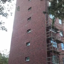 Wysoki budynek z czerwonej cegły z widocznymi rusztowaniami, częściowo zasłonięty przez gałęzie drzew, sugerujący prace elewacyjne w toku.