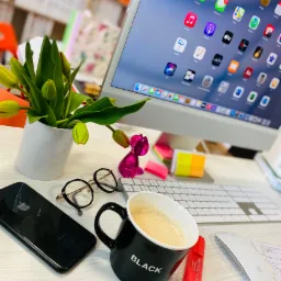 Biurko z iMaciem, czarnym telefonem iPhone, okularami, kubkiem z kawą z napisem BLACK, tulipanami w białym wazonie i różowym ozdobnym króliczkiem, tworzące przytulne miejsce pracy.