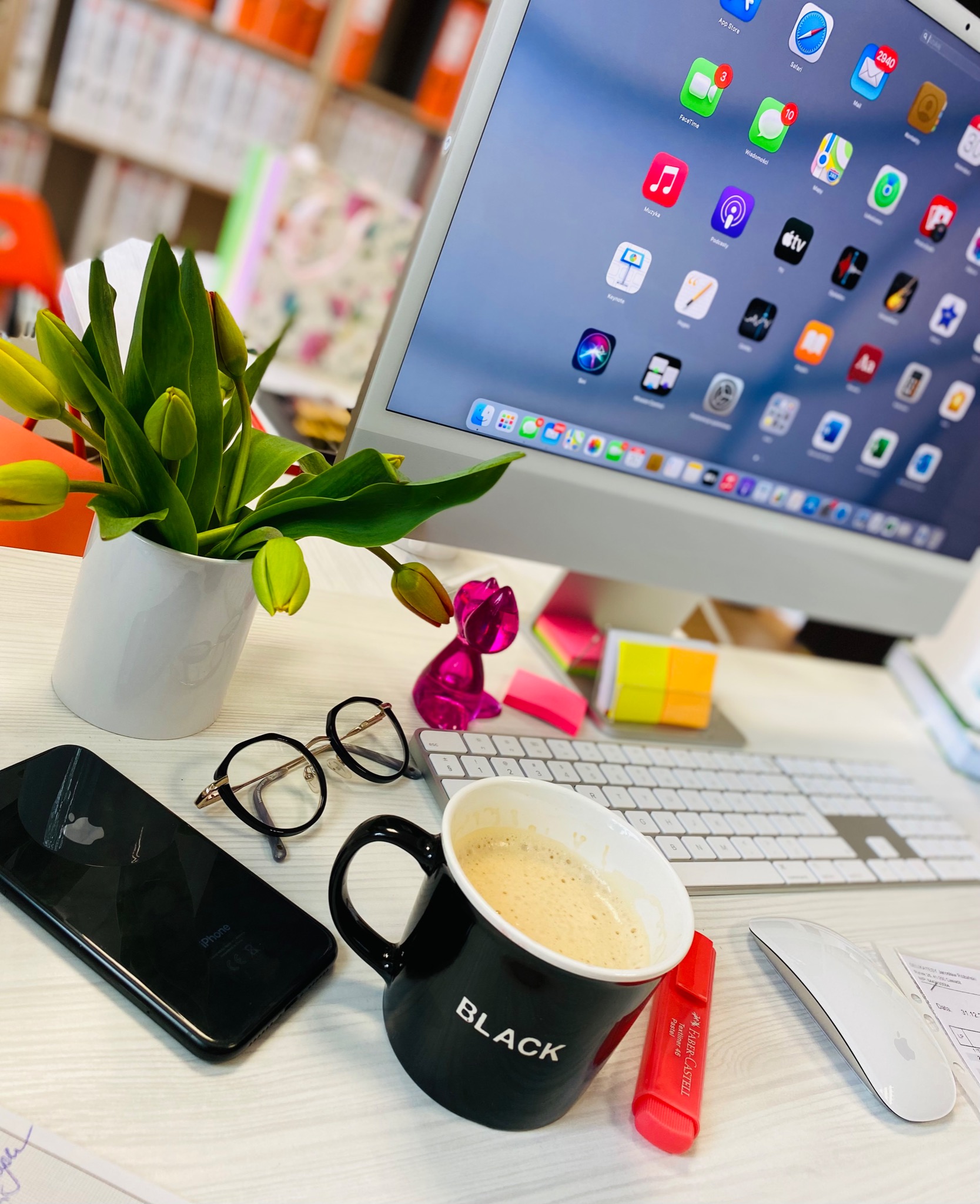 Biurko z iMaciem, czarnym telefonem iPhone, okularami, kubkiem z kawą z napisem BLACK, tulipanami w białym wazonie i różowym ozdobnym króliczkiem, tworzące przytulne miejsce pracy.