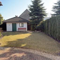 CZYSTY OGRÓD KAROL WIERZBICKI - Zadbany ogród z równo przystrzyżonym trawnikiem, żywopłotem z tui i małym domkiem narzędziowym z czerwoną ławeczką. Na dachu domku siedzi czarny kot.