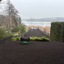 CZYSTY OGRÓD KAROL WIERZBICKI - Przygotowany teren pod trawnik z widokiem na jezioro. Świeżo nawieziona ziemia, widoczne krzewy i ogrodzenie. W tle woda i zalesione wzgórza w pochmurny dzień.