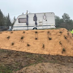 CZYSTY OGRÓD KAROL WIERZBICKI - Ukształtowany teren z agrowłókniną i sadzonkami, przyczepa kempingowa w tle, osoba w odblaskowej kamizelce robiąca zdjęcie. Prace ziemne i nasadzenia.