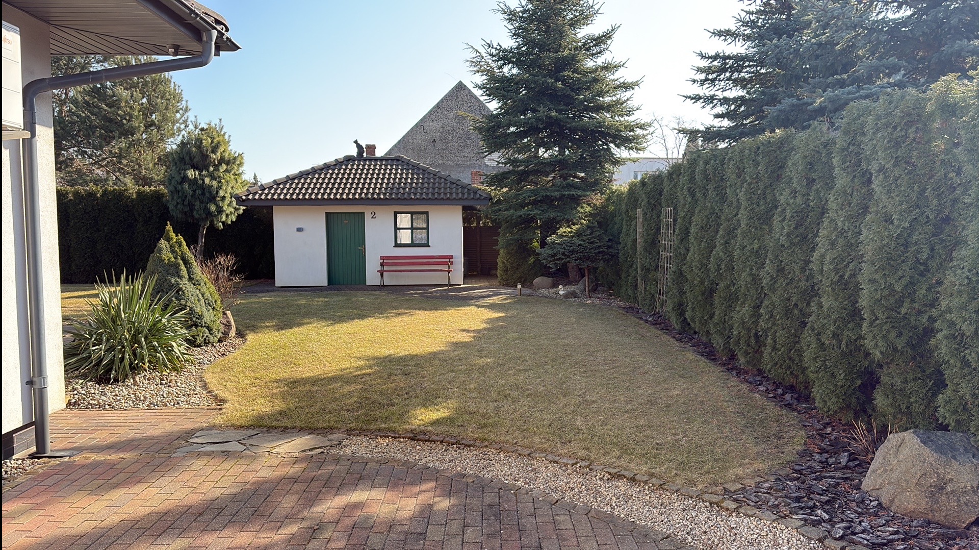Zadbany ogród z równo przystrzyżonym trawnikiem, żywopłotem z tui i małym domkiem narzędziowym z czerwoną ławeczką. Na dachu domku siedzi czarny kot.