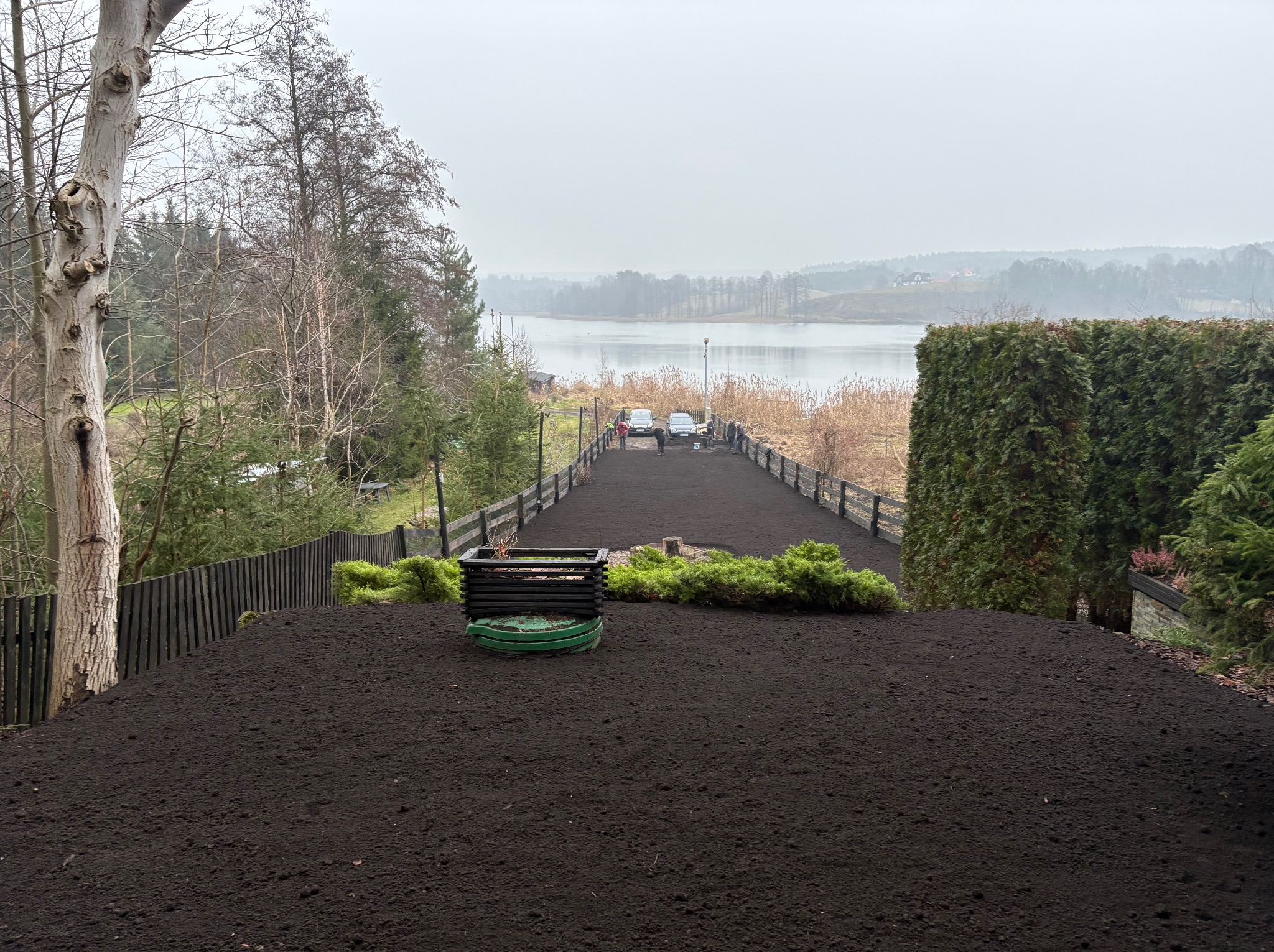 Przygotowany teren pod trawnik z widokiem na jezioro. Świeżo nawieziona ziemia, widoczne krzewy i ogrodzenie. W tle woda i zalesione wzgórza w pochmurny dzień.