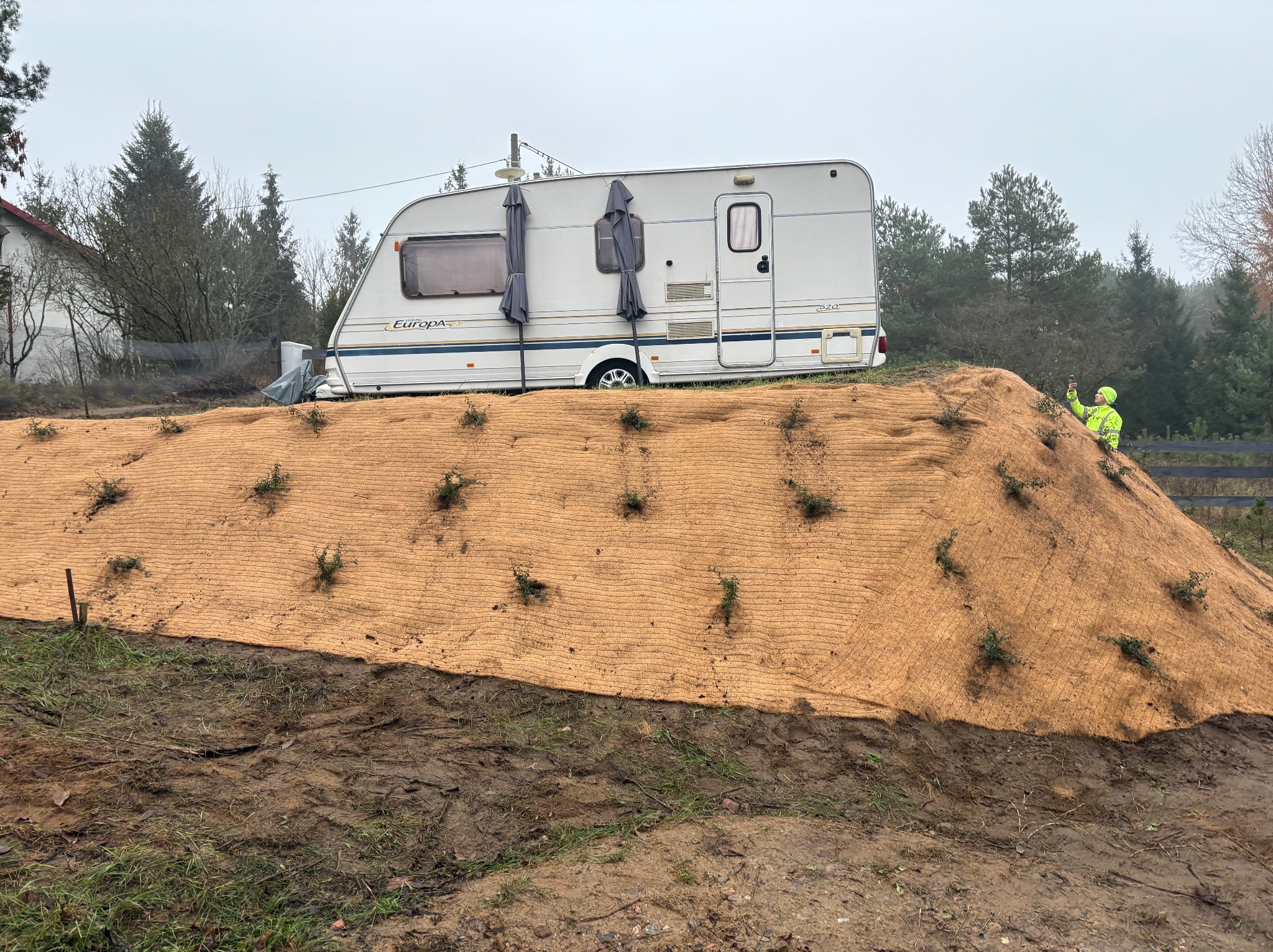 Ukształtowany teren z agrowłókniną i sadzonkami, przyczepa kempingowa w tle, osoba w odblaskowej kamizelce robiąca zdjęcie. Prace ziemne i nasadzenia.