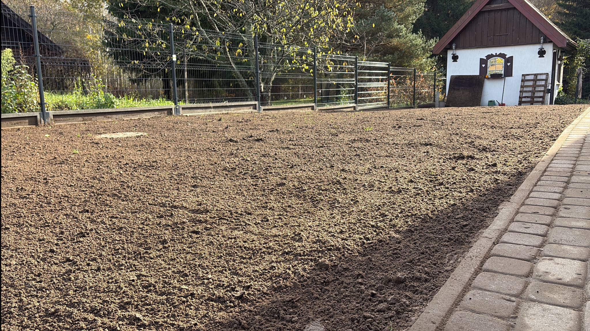 Przygotowany teren pod trawnik z widoczną ziemią, ogrodzeniem i budynkiem gospodarczym w tle. Świeżo przekopana gleba czeka na zasiew trawy.