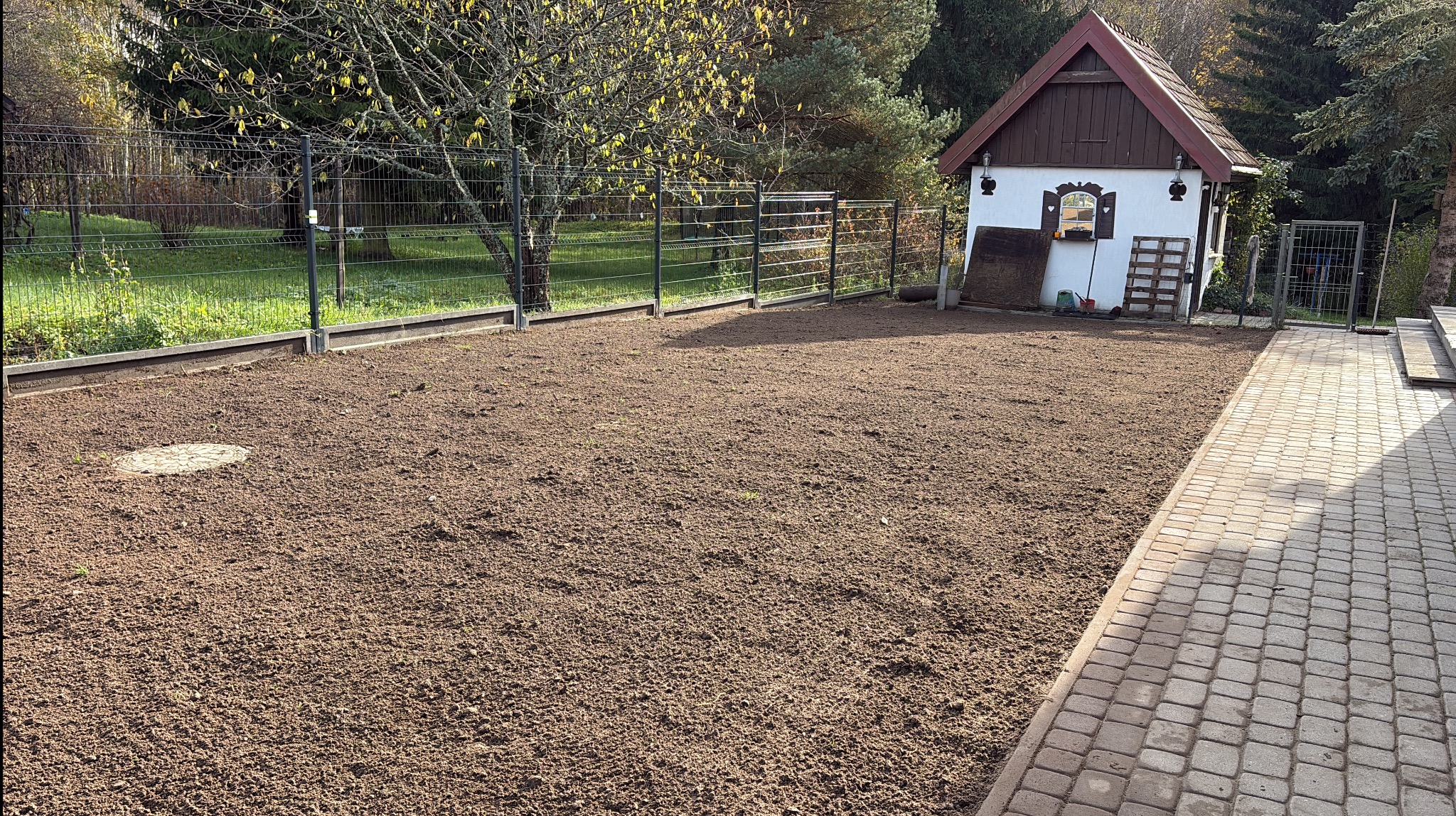 Przygotowany teren pod trawnik z widoczną ziemią, ogrodzeniem i małym budynkiem gospodarczym w tle. Etap prac ziemnych przed siewem trawy.