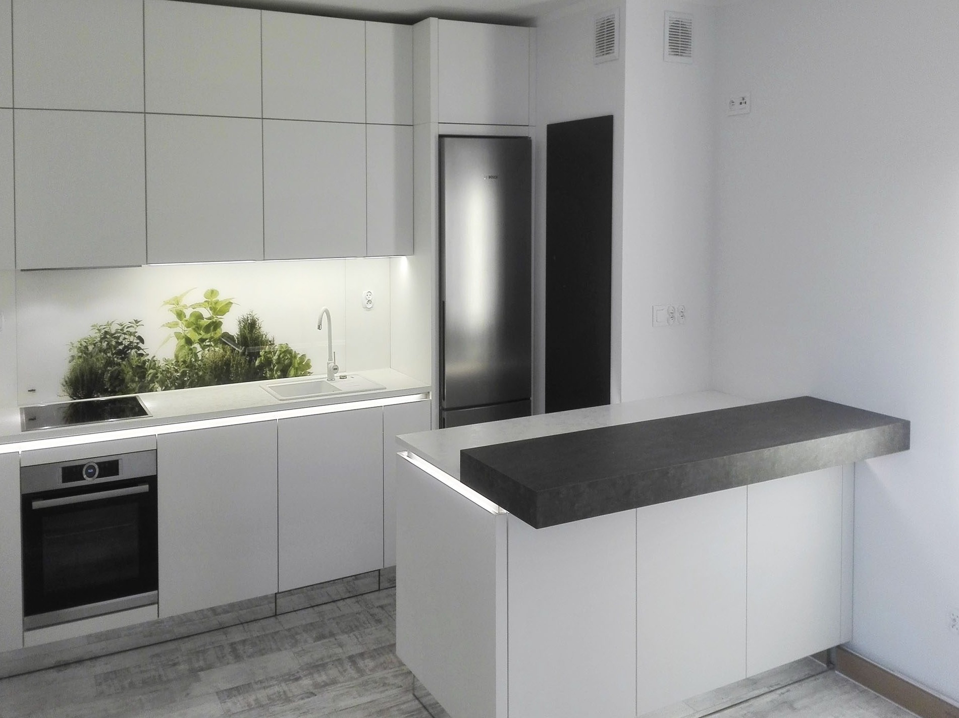 Nowoczesna, minimalistyczna kuchnia z białymi frontami, szarym blatem wyspy i panelem ze świeżymi ziołami nad zlewem.