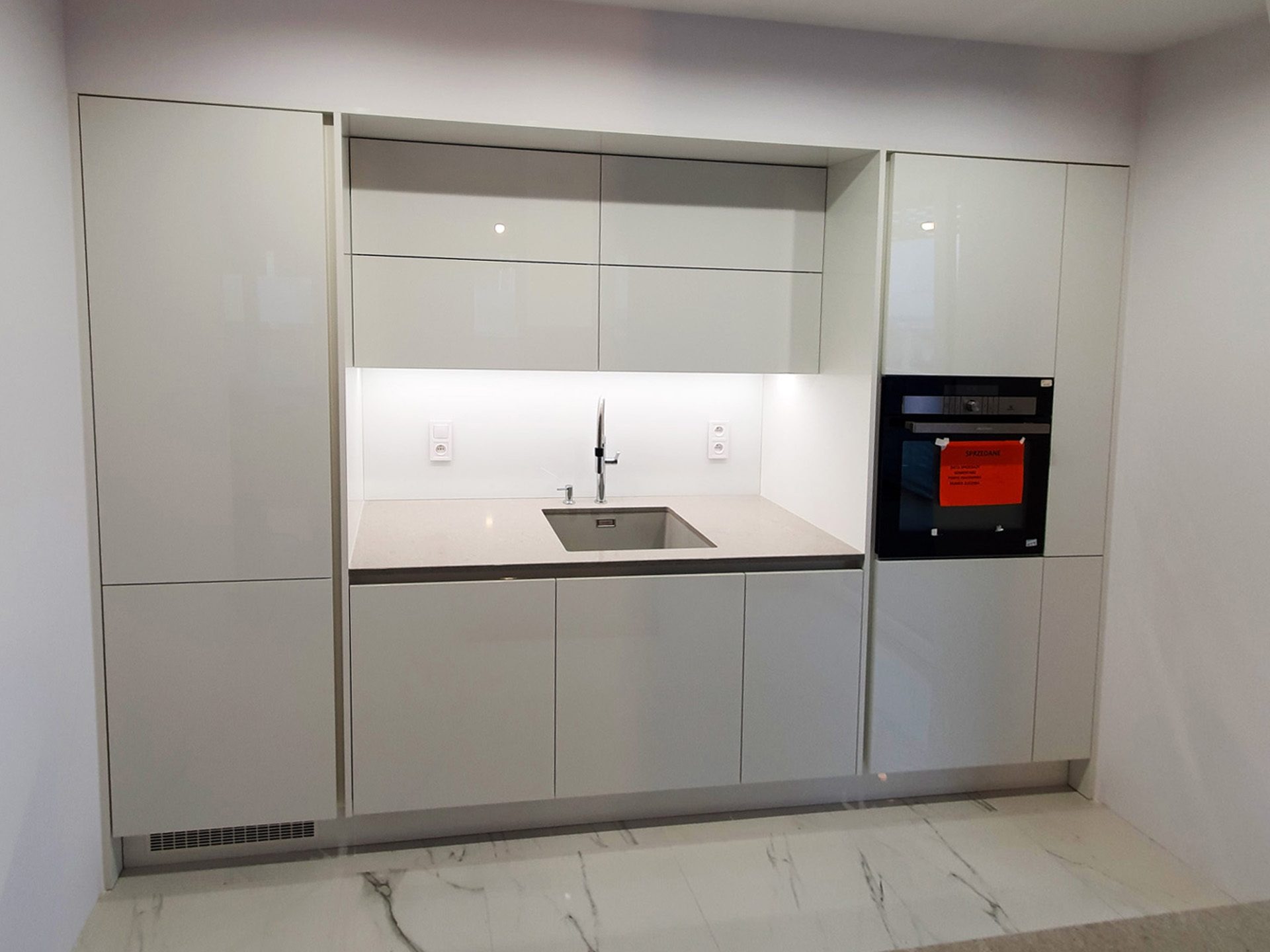 Nowoczesna, minimalistyczna kuchnia na wymiar we Wrocławiu z białymi, lakierowanymi frontami, szarym blatem i marmurową podłogą. W zabudowie piekarnik z czerwoną naklejką informacyjną.