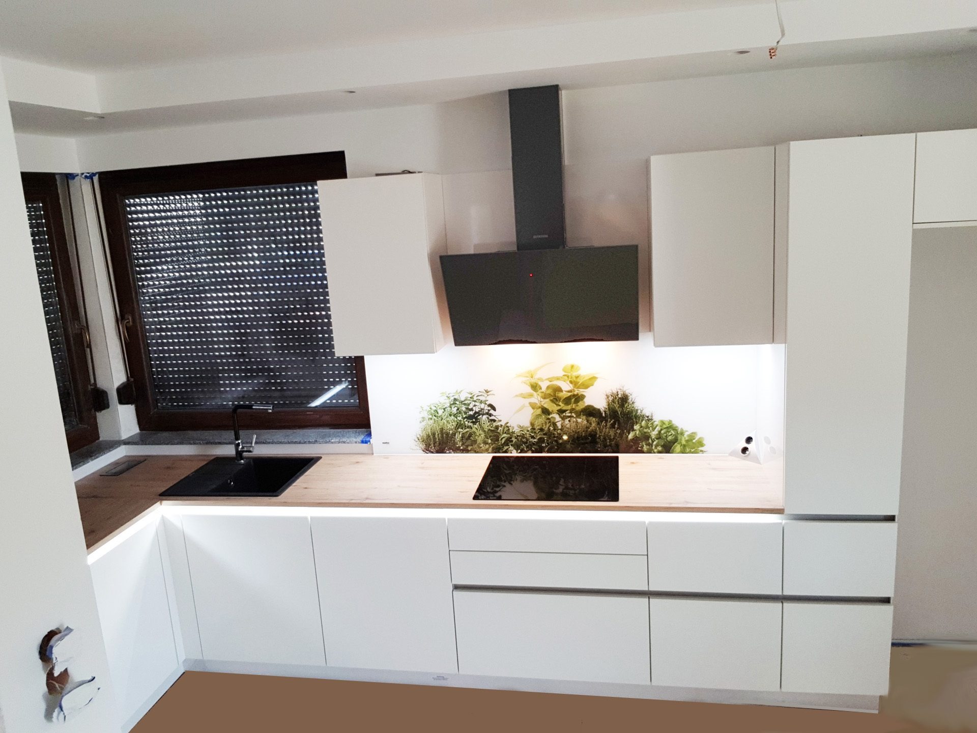 Nowoczesna, biała zabudowa kuchenna z drewnianym blatem, czarnym zlewem i płytą indukcyjną, oraz panelem szklanym z motywem ziół nad blatem, pod czarnym okapem.