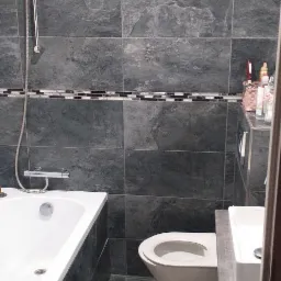 Wykończona łazienka z szarymi płytkami imitującymi kamień, białą wanną, toaletą i umywalką. Dekoracyjny pasek z mozaiki na ścianie. Beżowy dywanik na podłodze.