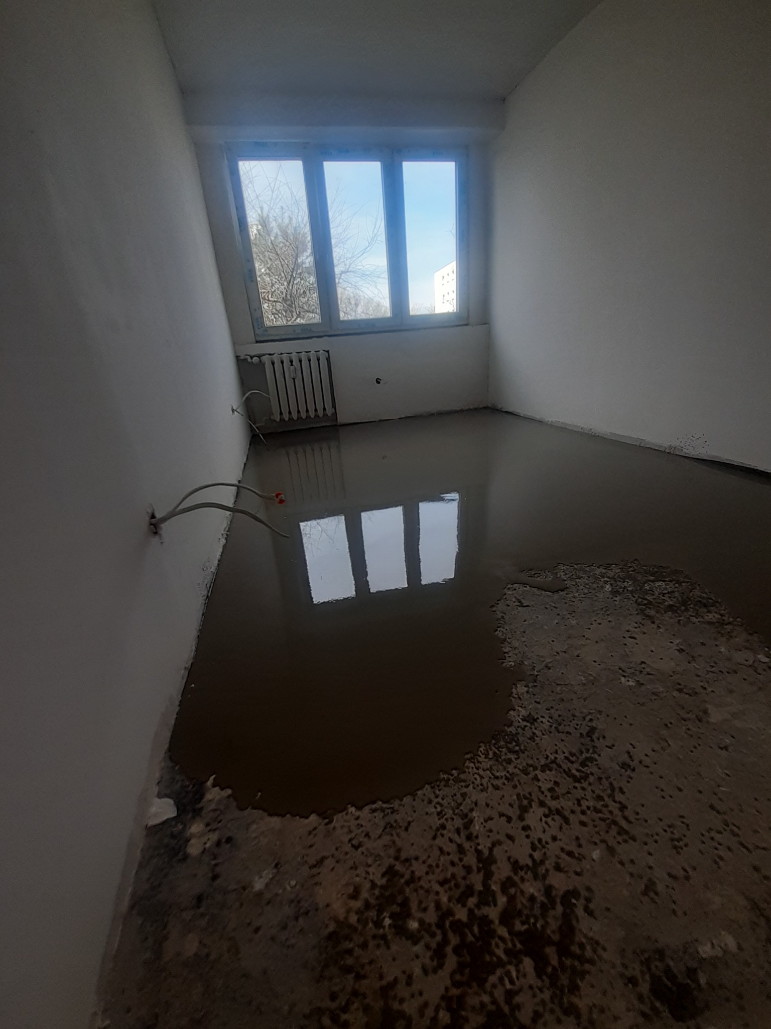 Pokój w trakcie wylewania posadzki, widoczne kable wystające ze ściany i okno z widokiem na drzewo.