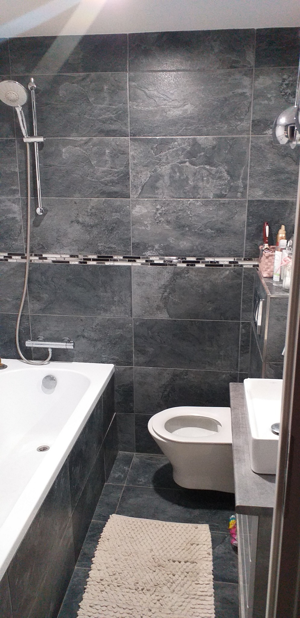 Wykończona łazienka z szarymi płytkami imitującymi kamień, białą wanną, toaletą i umywalką. Dekoracyjny pasek z mozaiki na ścianie. Beżowy dywanik na podłodze.