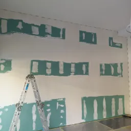 Ściana po szpachlowaniu, z widocznymi łatami szpachli w kolorze miętowym, przygotowana do dalszych prac wykończeniowych. Aluminiowa drabina rozstawiona przy ścianie, podłoga zabezpieczona folią...