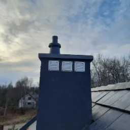 Ciemny komin wentylacyjny z trzema metalowymi kratkami na dachu pokrytym łupkiem, na tle pochmurnego nieba i fragmentu zabudowań.