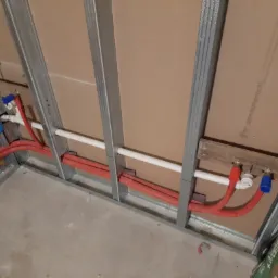 Instalacja rur grzewczych i wodnych w ścianie szkieletowej z płyt gipsowo-kartonowych w trakcie prac instalacyjnych w Klecza Górna, widoczne czerwone rury PEX i białe rury PCV, niebieskie zawory...