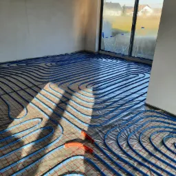 Niebieskie rury ogrzewania podłogowego ułożone na siatce w surowym pomieszczeniu z dużym oknem, widoczne pomarańczowe złącza, w tle zabudowa jednorodzinna.