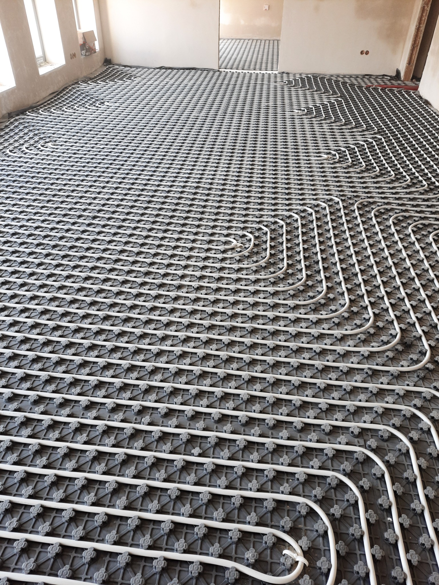 Rozłożona instalacja ogrzewania podłogowego na czarnej macie izolacyjnej, widoczne rury w kształcie pętli, surowy stan wykończenia pomieszczenia.