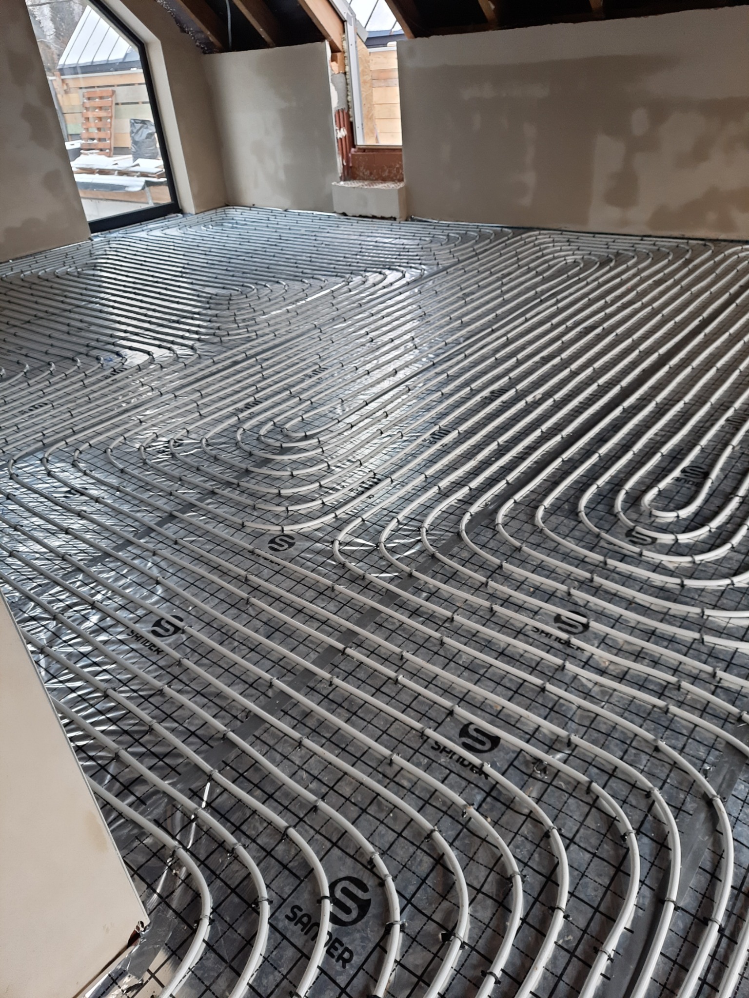 Białe rurki ogrzewania podłogowego ułożone w pętle na czarnej siatce, pokrytej folią izolacyjną z nadrukowanym logo producenta, widok na surową podłogę w trakcie remontu.