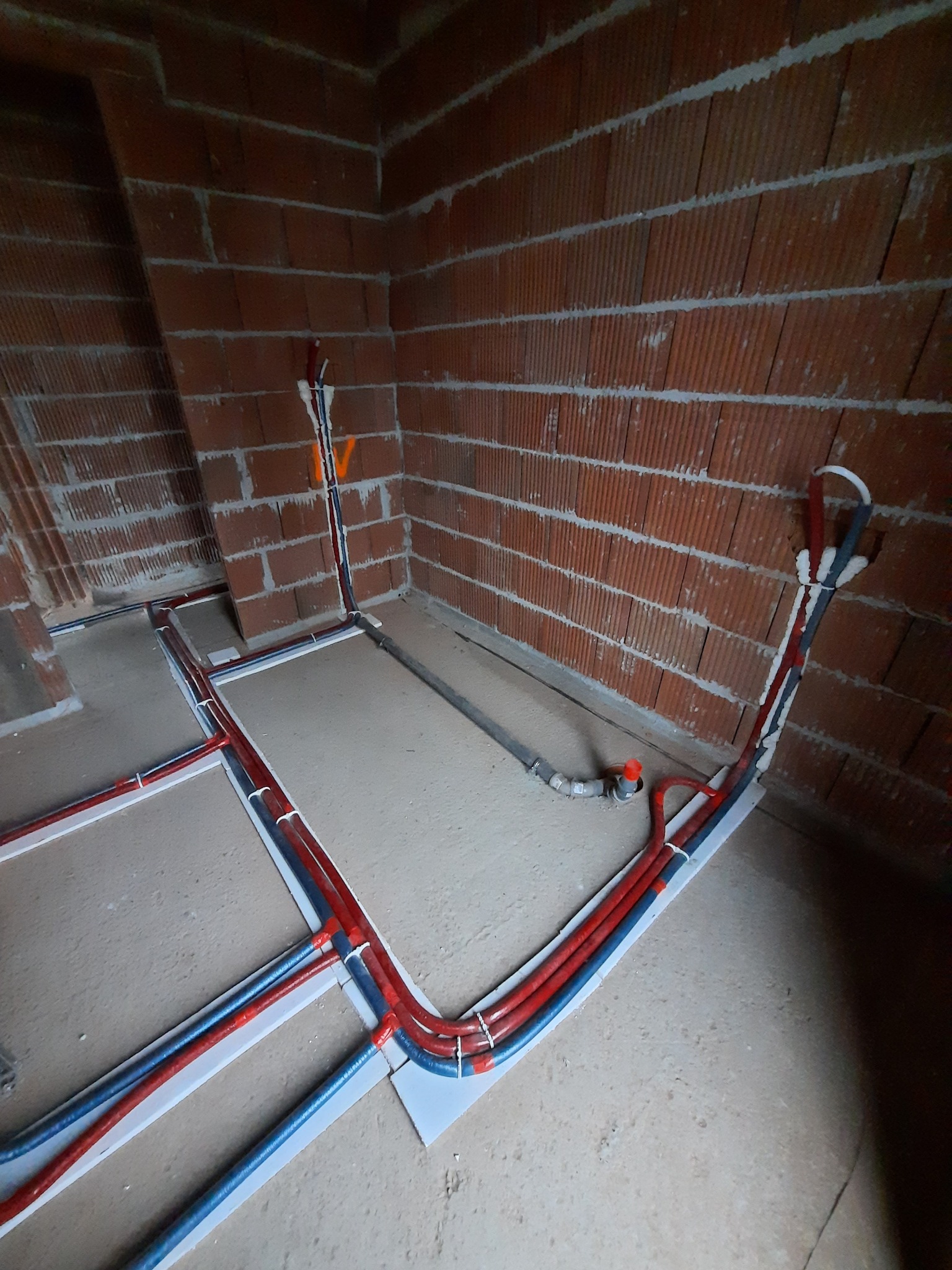 Instalacja ogrzewania podłogowego z rurami w kolorze czerwonym i niebieskim, przymocowanymi do podłoża listwami, widok z góry w surowym wnętrzu z ceglanymi ścianami.