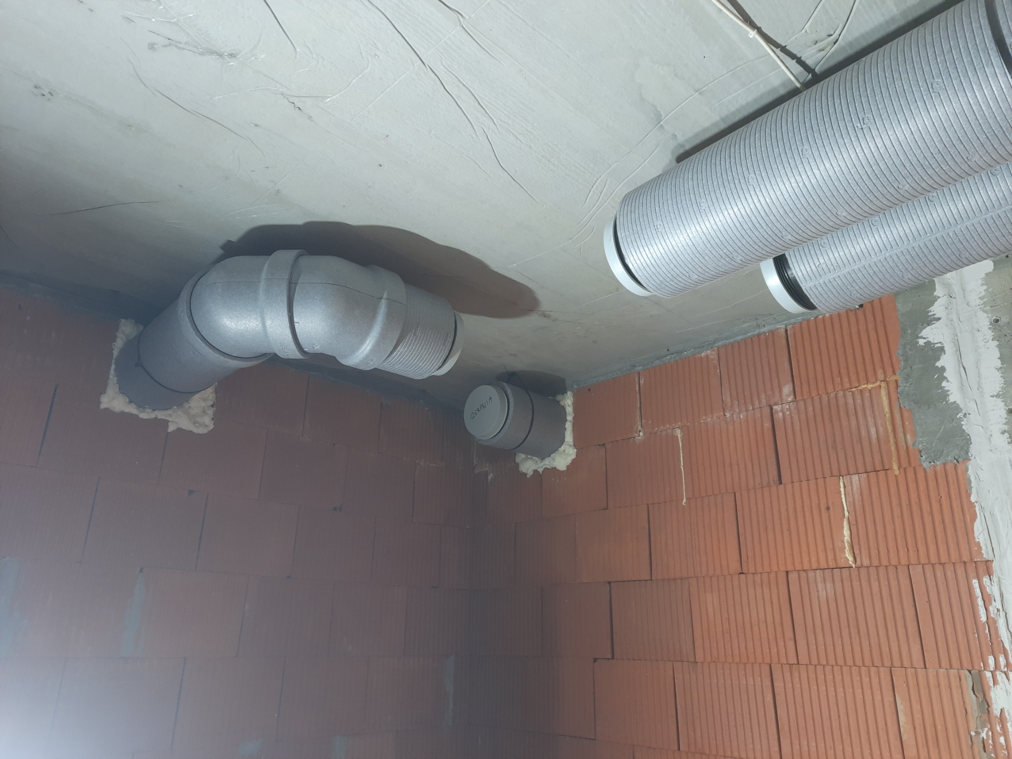 Szare rury wentylacyjne przymocowane do sufitu i ściany z czerwonej cegły, uszczelnione pianką montażową.