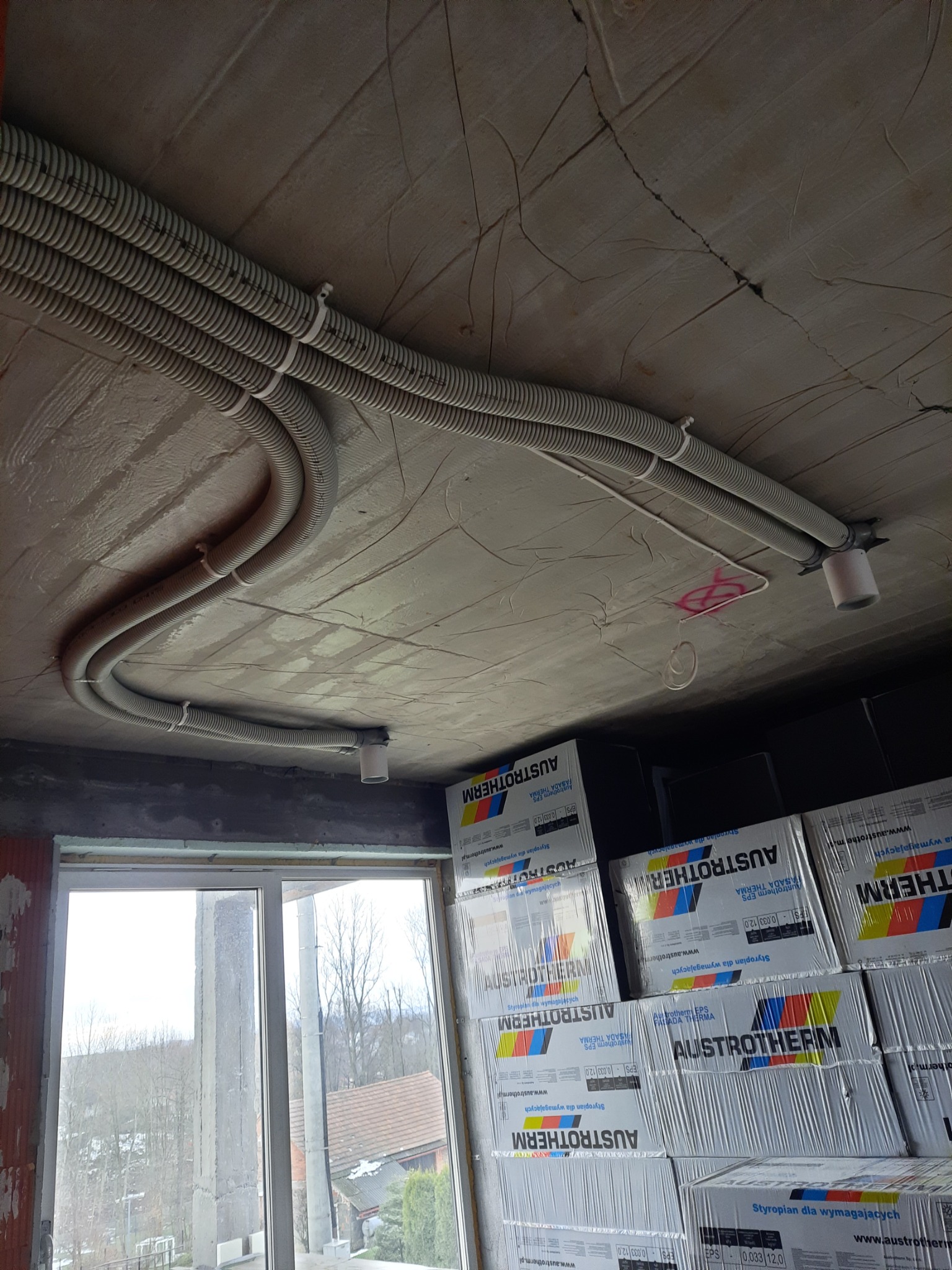 Montaż peszli instalacyjnych na suficie w surowym stanie, widoczne uchwyty mocujące i puszki instalacyjne, w tle okno i stos paczek styropianu Austrotherm.