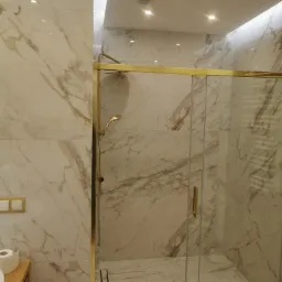 Kabina prysznicowa z marmurowymi płytkami w odcieniach bieli i złota, widoczny odpływ liniowy i armatura w kolorze złotym.