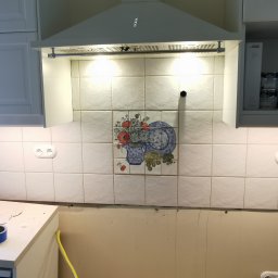 Dawid Baran Remonty i wykonczenis - Fragment kuchni w trakcie remontu: białe płytki z dekoracyjnym motywem, widoczne gniazdka elektryczne, okap i fragment szafek. Taśma malarska zabezpiecza krawędzie.