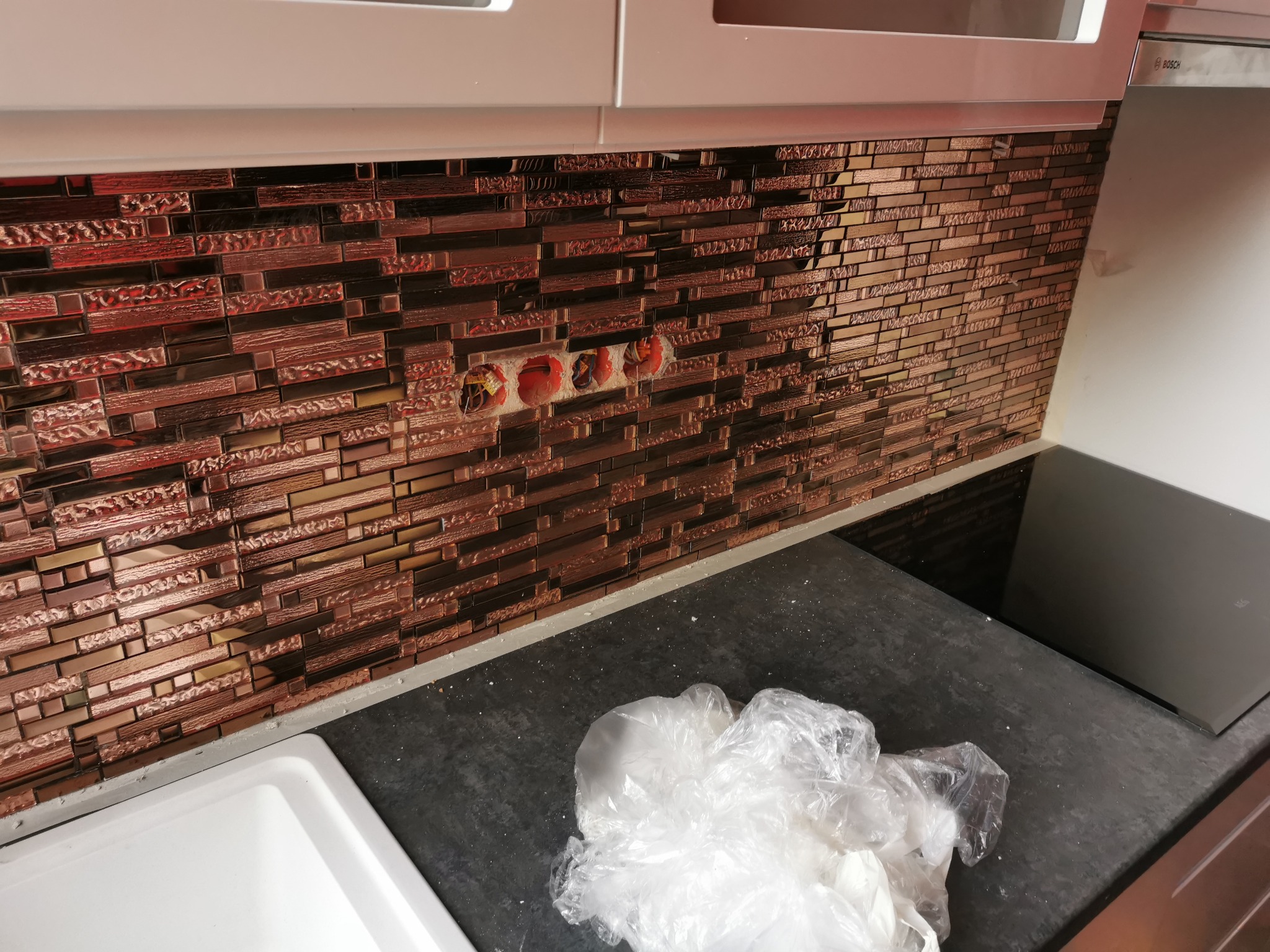 Błyszcząca mozaika w odcieniach brązu i miedzi na ścianie w kuchni, z widocznymi otworami na gniazdka elektryczne i fragmentem blatu oraz zlewu.