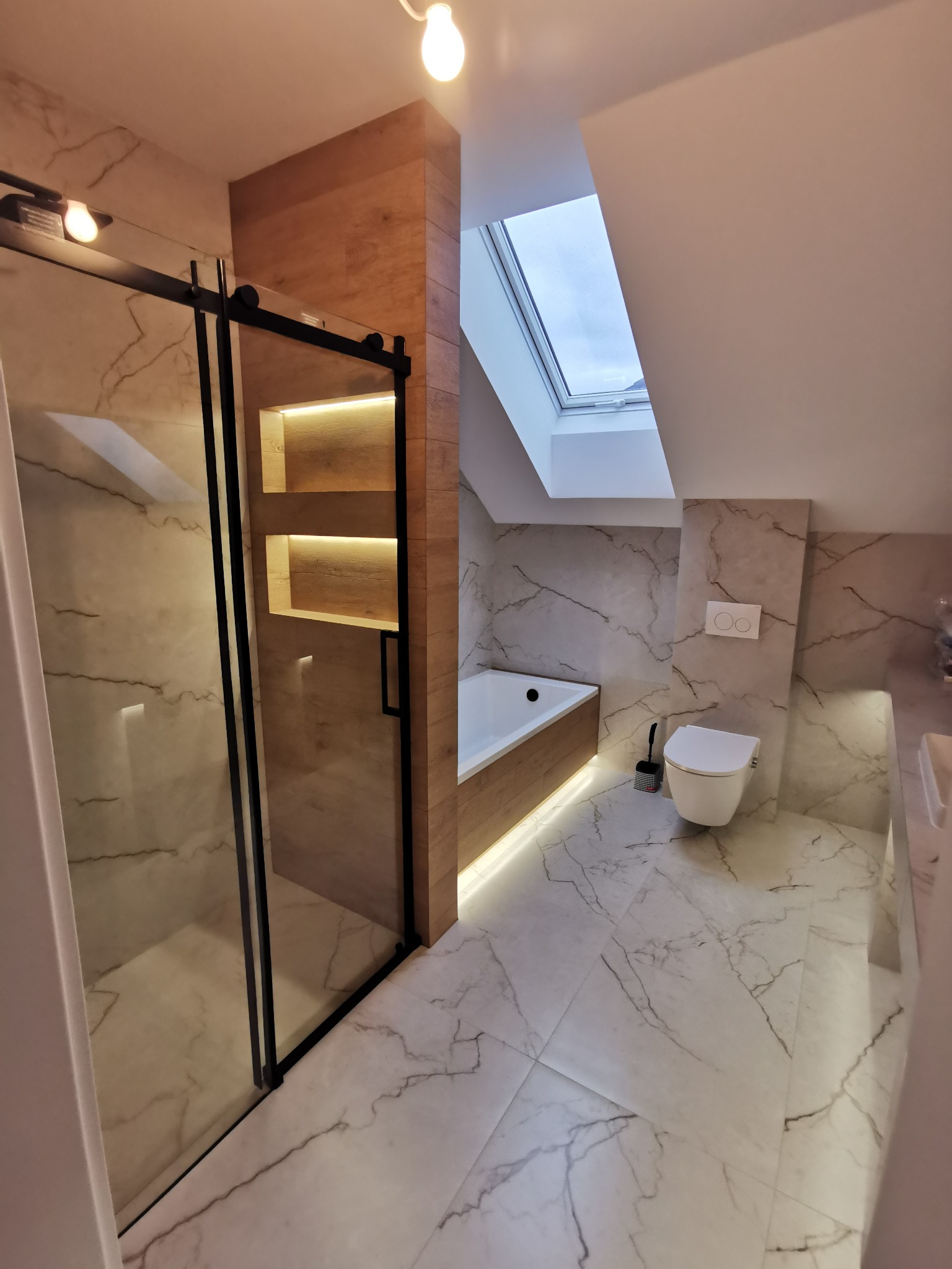 Nowoczesna łazienka na poddaszu z marmurowymi płytkami, kabiną prysznicową typu walk-in z czarnymi ramami i drewnianymi akcentami. Oświetlenie LED podkreśla detale.