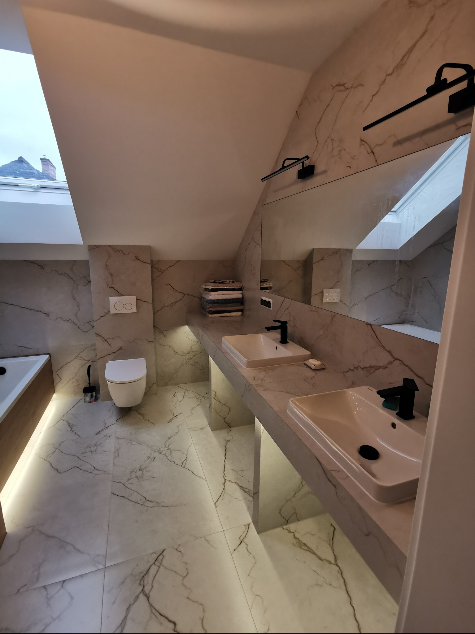 Nowoczesna łazienka na poddaszu z marmurowymi płytkami, podświetleniem LED pod szafką umywalkową i dużym lustrem. Widoczny fragment okna dachowego.