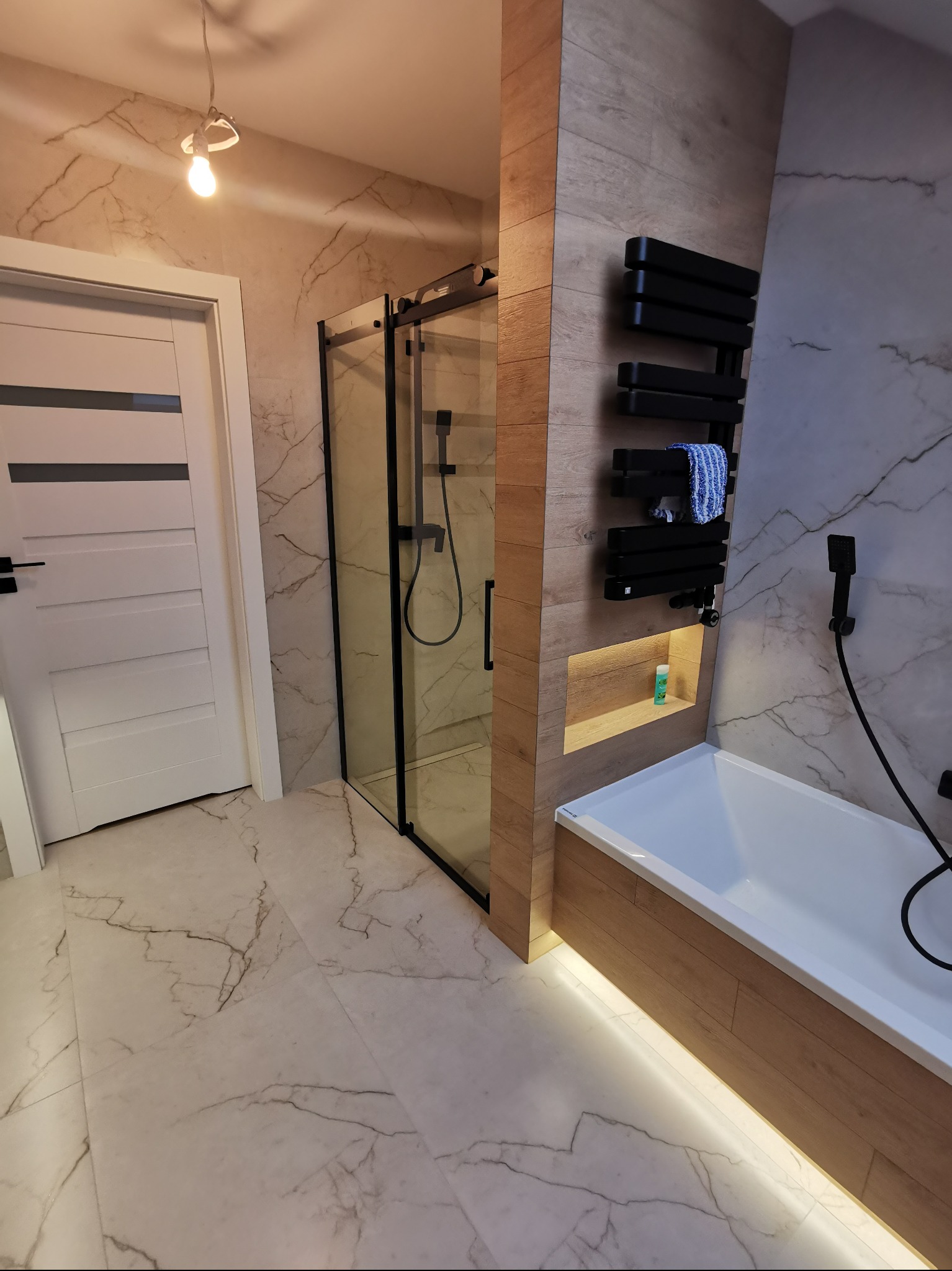 Nowoczesna łazienka z kabiną prysznicową typu walk-in, grzejnikiem drabinkowym i podświetlaną wanną. Płytki imitujące marmur na podłodze i ścianach.