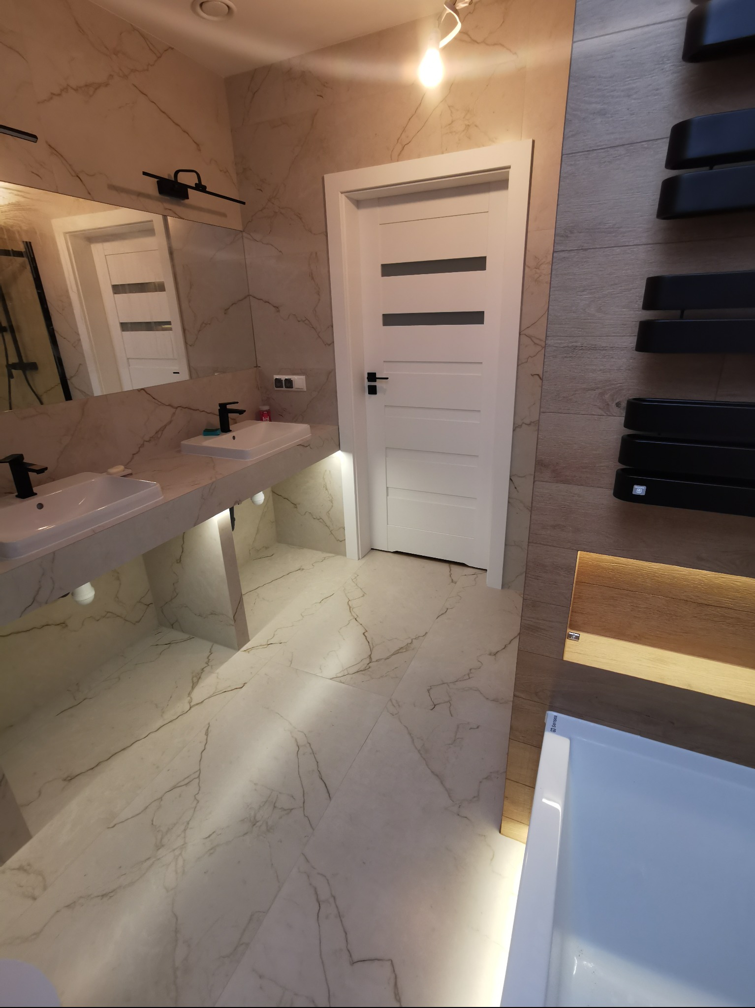 Nowoczesna łazienka w Czernichowie z marmurowymi płytkami, podświetlanym blatem z umywalkami i grzejnikiem drabinkowym. Białe drzwi z poziomymi frezami.