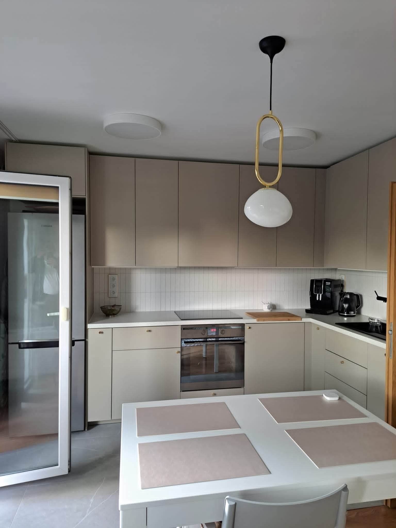 Jasna kuchnia w stylu minimalistycznym z beżowymi szafkami, białymi płytkami nad blatem i nowoczesnym oświetleniem. Widoczny stół z nakryciami i sprzęt AGD.