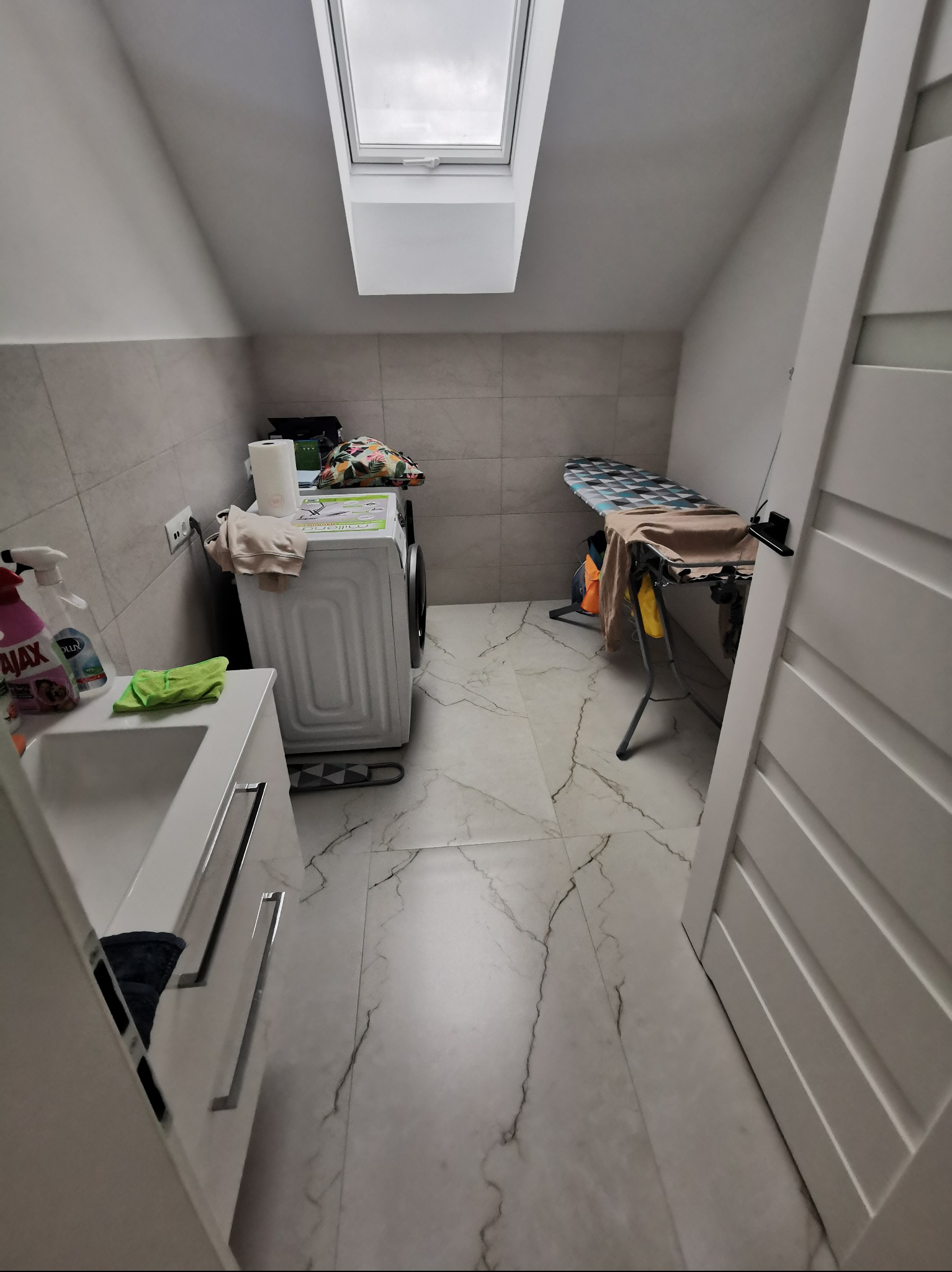 Jasna łazienka na poddaszu z pralką, umywalką z szafką, deską do prasowania i oknem dachowym. Płytki imitujące marmur na podłodze i ścianie.