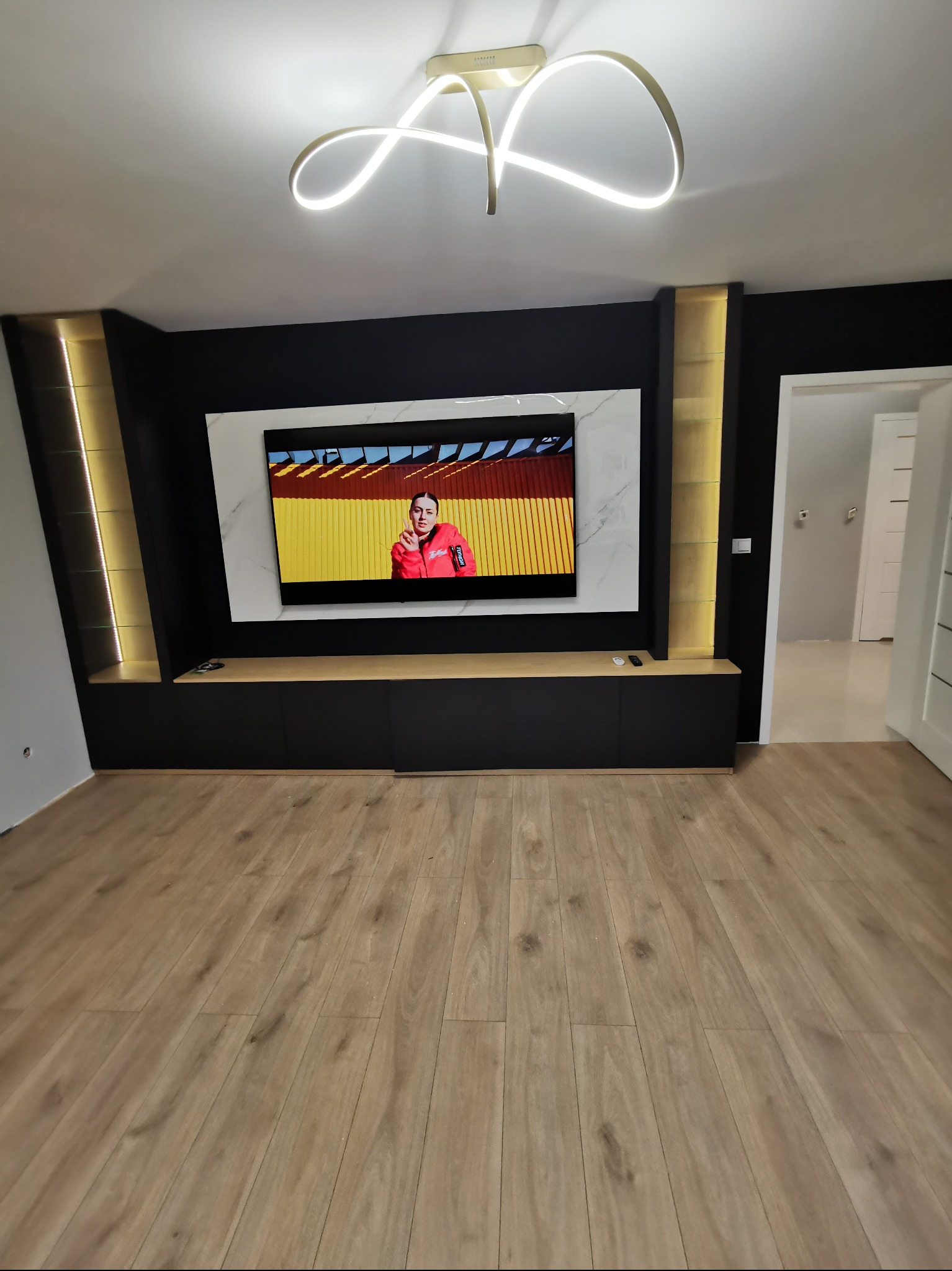 Nowoczesny salon z czarną zabudową TV, marmurowym panelem i podświetlanymi półkami. Parkiet w odcieniach drewna. Sufit zdobi designerska lampa LED.