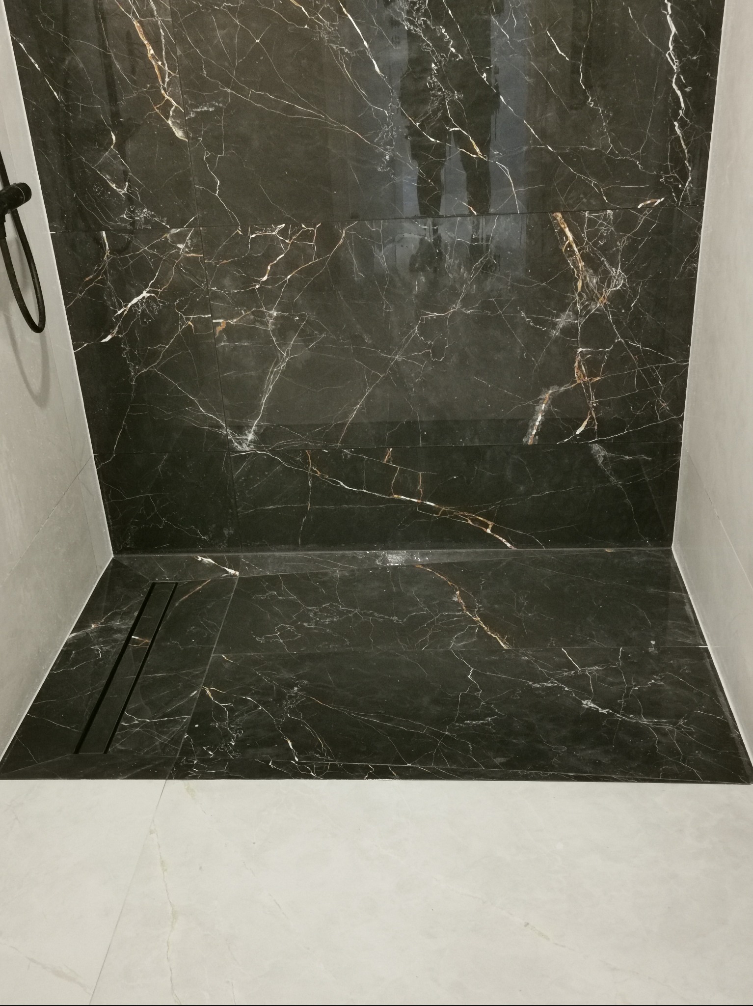 Wykończona kabina prysznicowa z czarnymi płytkami imitującymi marmur z białymi i złotymi żyłami, z odpływem liniowym i szarą ścianą boczną.