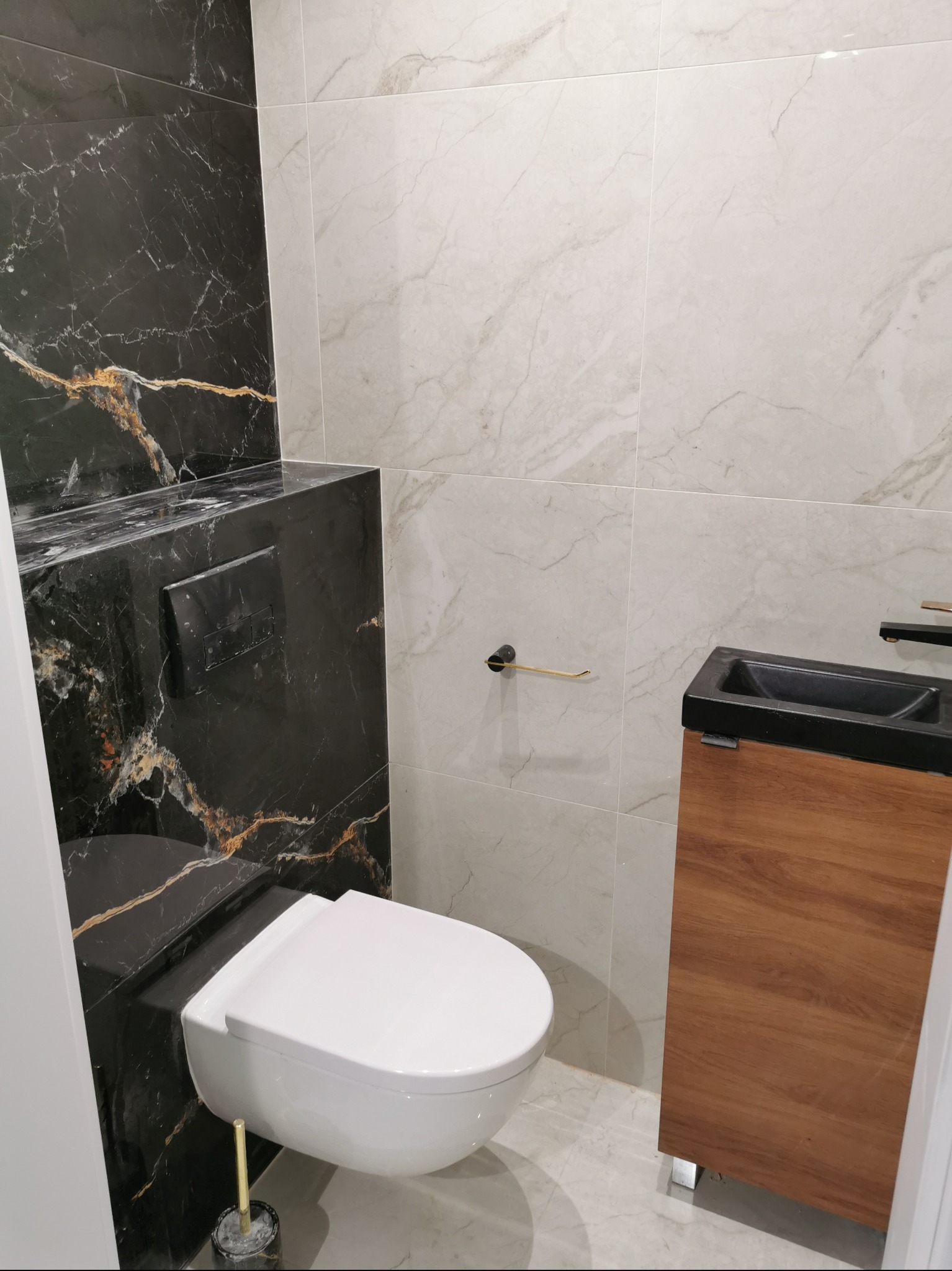 Nowoczesna toaleta z marmurowymi płytkami w odcieniach bieli i czerni ze złotymi akcentami, wisząca muszla klozetowa i umywalka z szafką z drewnopodobnym wykończeniem.