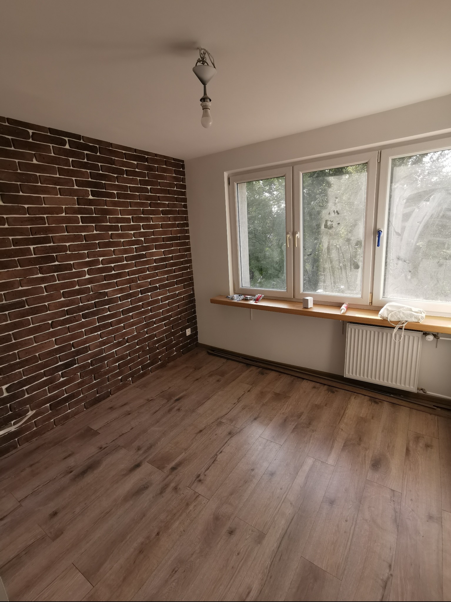 Pokój po remoncie: ściana z cegły dekoracyjnej, panele podłogowe, okno z drewnianym parapetem, grzejnik. Widok na wnętrze z surową żarówką.