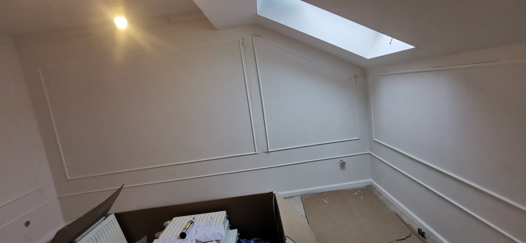 Białe ściany z dekoracyjnymi listwami w pokoju z oknem dachowym. Karton z elementami grzejnika i planami na podłodze. Widok na wykończone wnętrze.