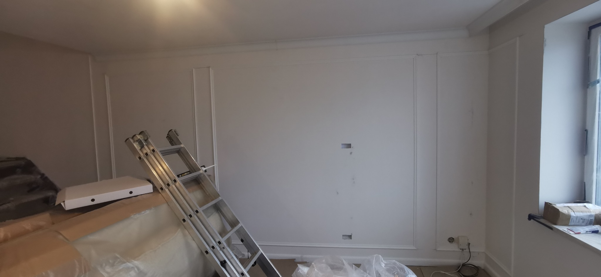 Białe ściany z dekoracyjnymi listwami w trakcie wykańczania. Widoczna drabina, zabezpieczone folią meble i przygotowane otwory na gniazdka elektryczne.