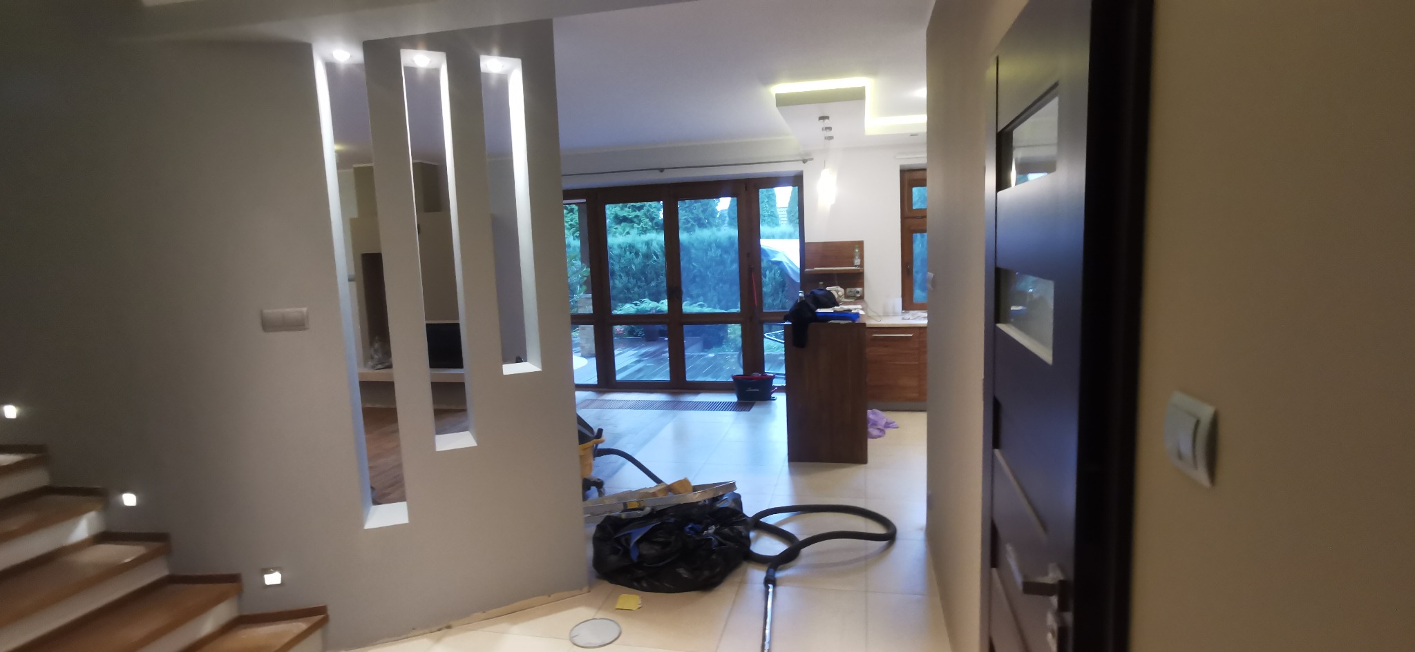 Nowoczesne wnętrze domu z widokiem na ogród przez duże okna. Elementy dekoracyjne z podświetleniem. Widoczne schody i fragment wyposażenia kuchni.