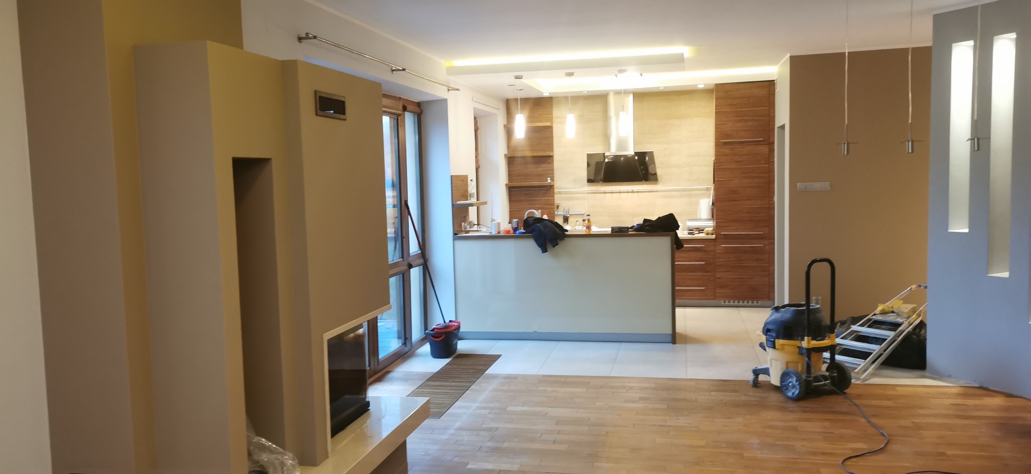 Jasne, przestronne wnętrze po remoncie z drewnianą podłogą, nowoczesnym kominkiem i widoczną kuchnią. Sprzęt budowlany sugeruje prace wykończeniowe.