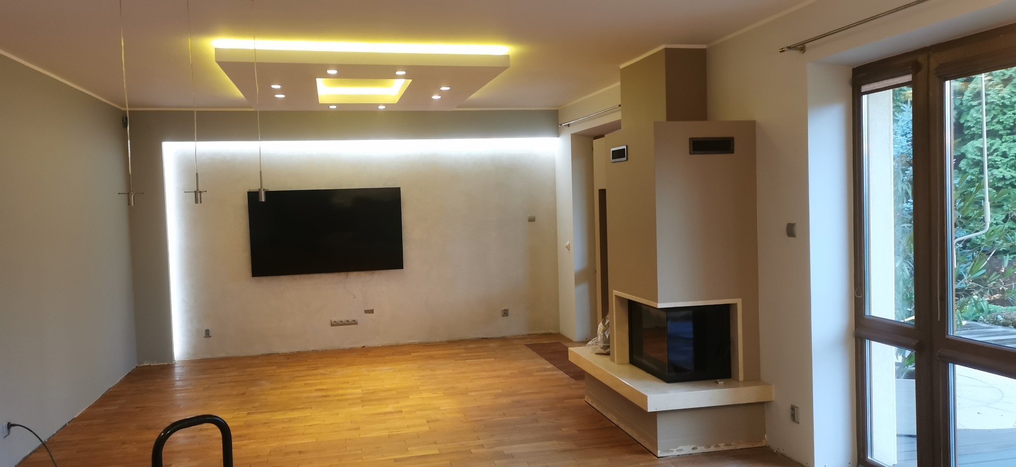 Jasne wnętrze po remoncie z drewnianą podłogą, nowoczesnym kominkiem i podwieszanym sufitem z oświetleniem LED. Widoczne gładkie ściany i duże okno z widokiem na zieleń.