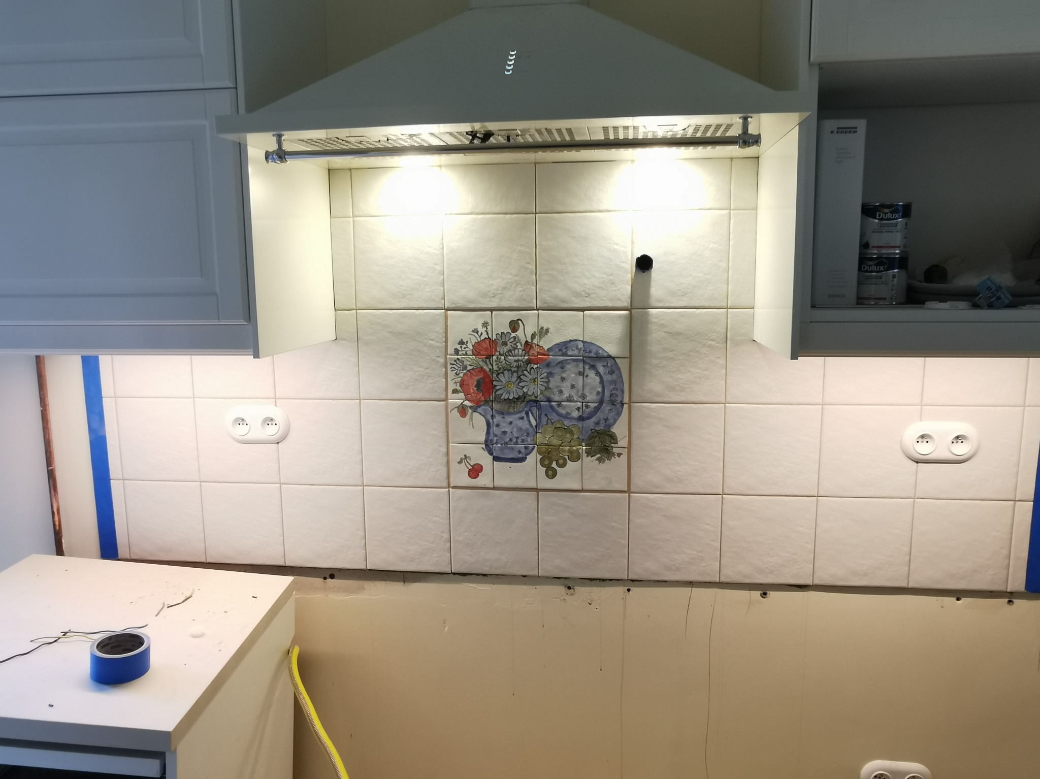 Fragment kuchni w trakcie remontu: białe płytki z dekoracyjnym motywem, widoczne gniazdka elektryczne, okap i fragment szafek. Taśma malarska zabezpiecza krawędzie.